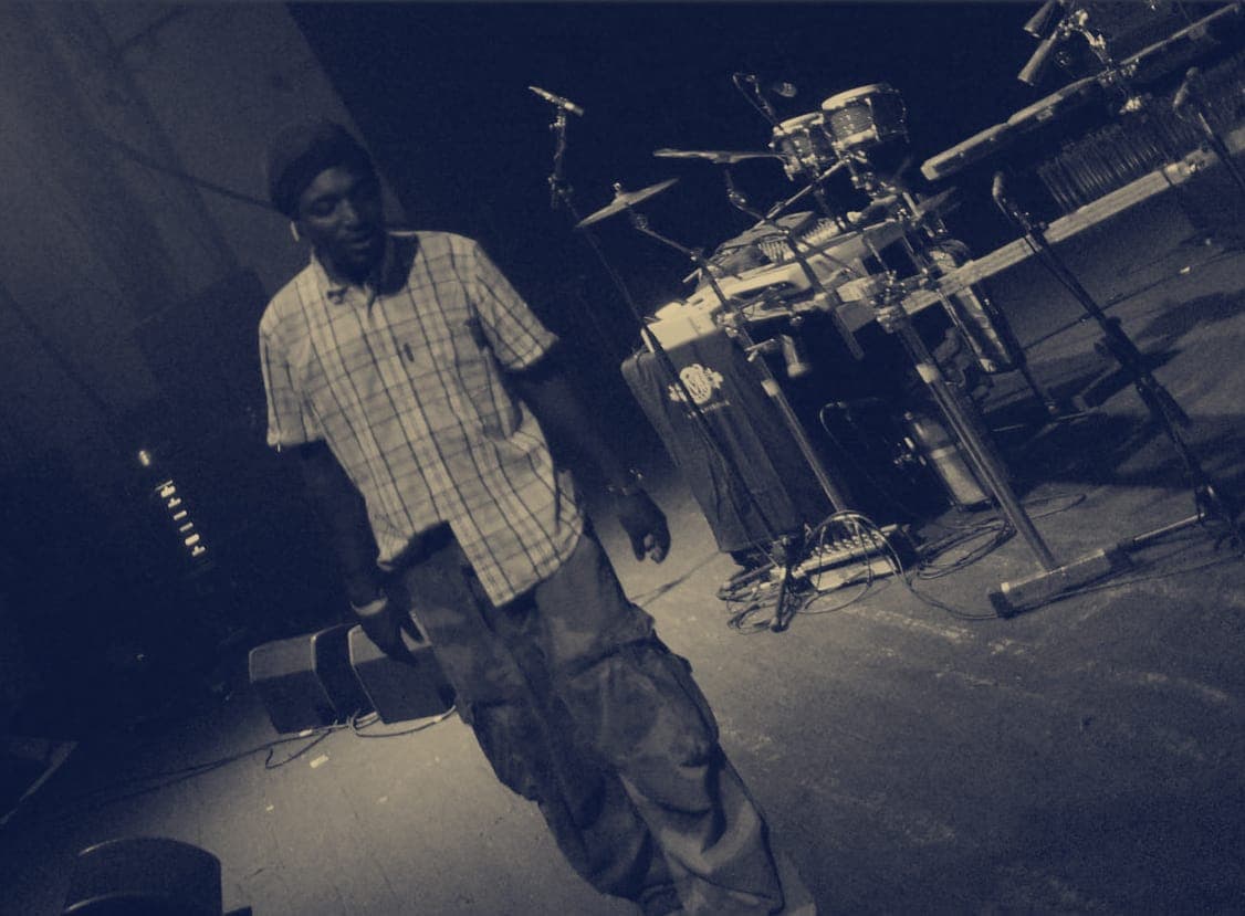 Kareem Shabazz walking the stage at Brixton Academy in a checked shirt, the drum kit behind him