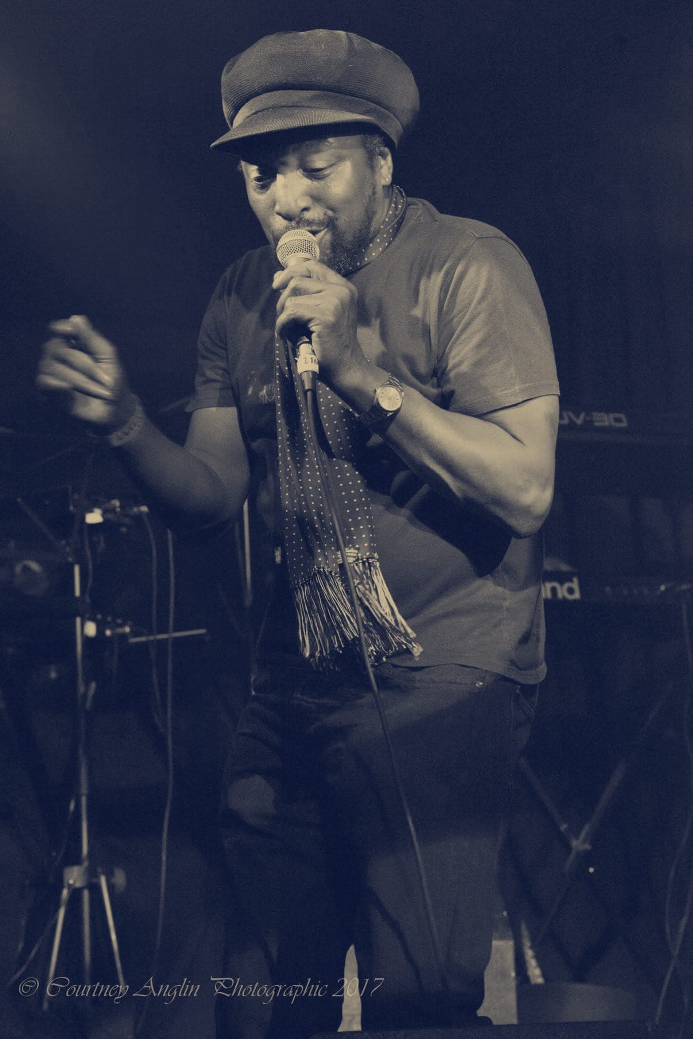 Kareem Shabazz singing under green stage light in a flat cap and scarf