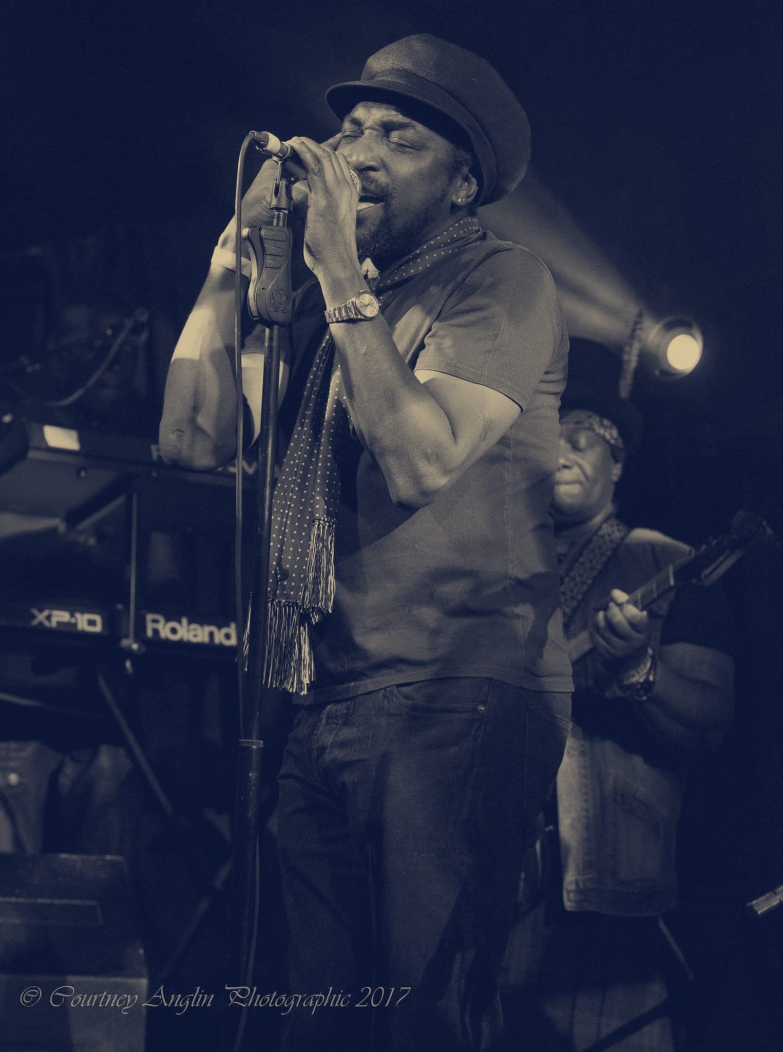 Kareem Shabazz singing at a microphone stand in front of a keyboard, a scarf over his shoulders