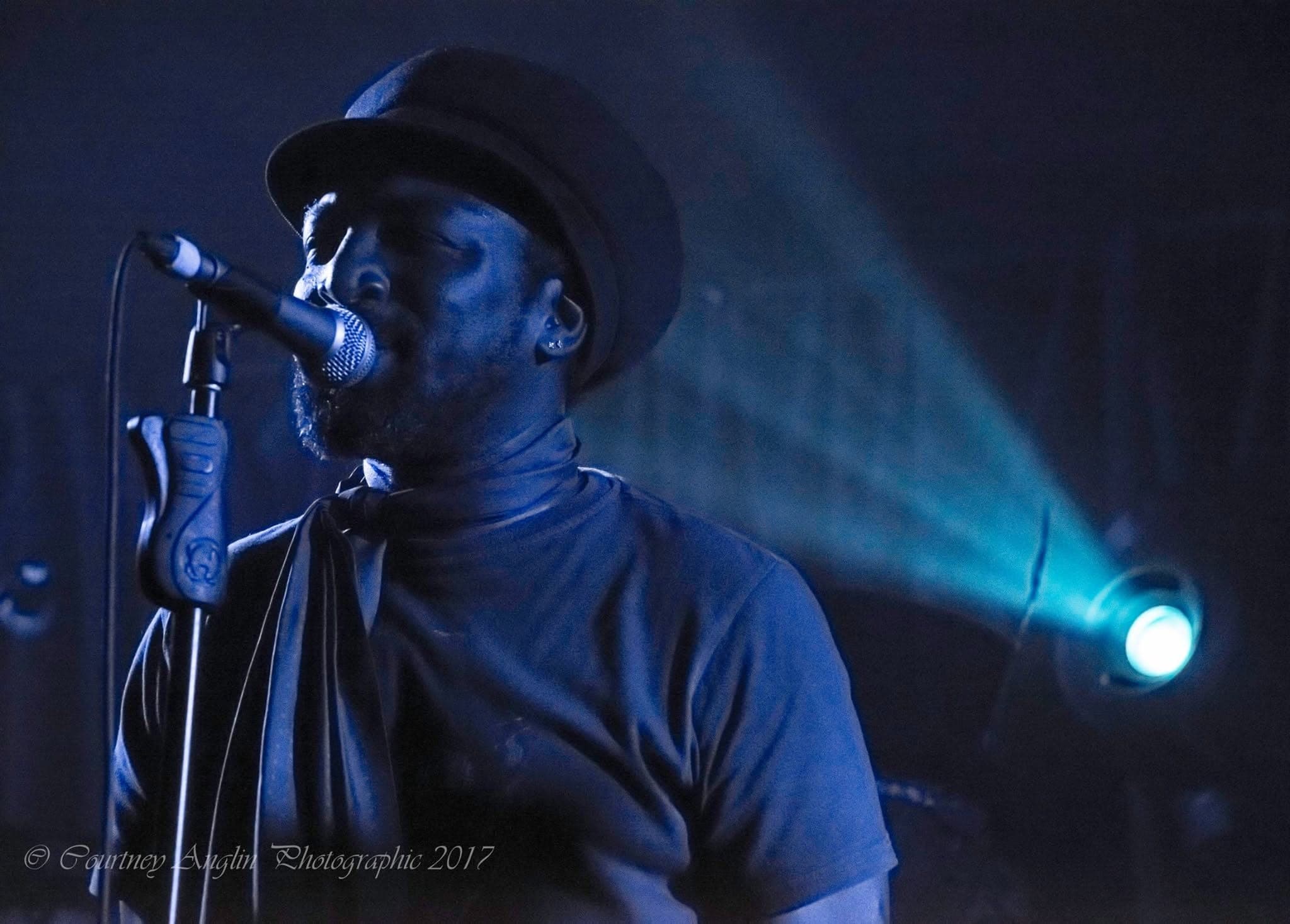 Kareem Shabazz singing at a microphone stand, a stage light beam cutting across behind him