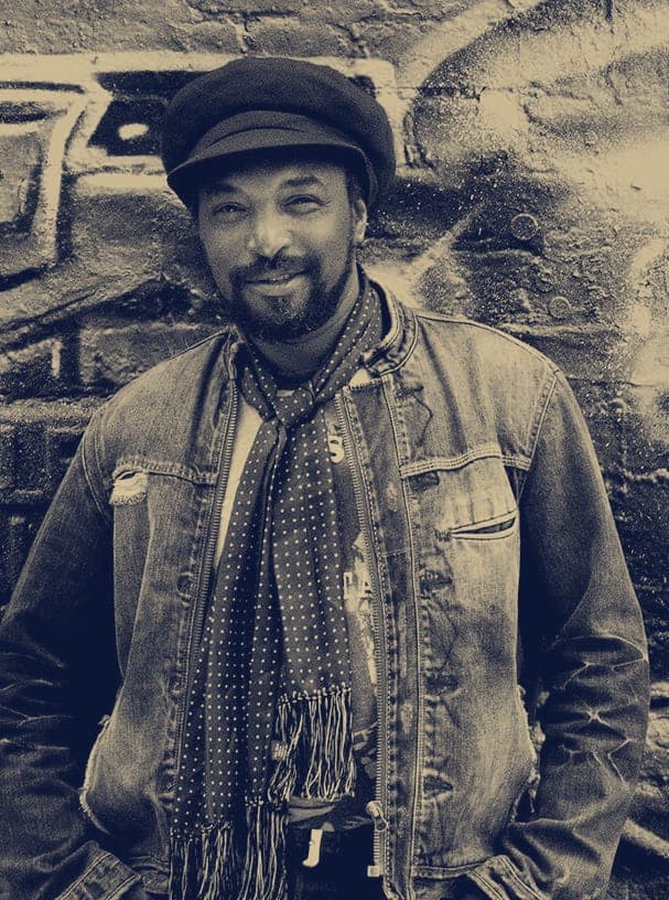 Kareem Shabazz standing in a denim jacket and scarves against a graffiti wall