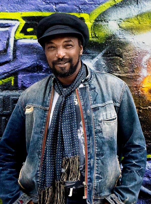 Kareem Shabazz standing in a denim jacket and scarves against a graffiti wall