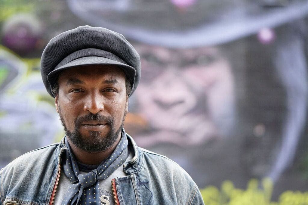Portrait of Kareem Shabazz in a flat cap and denim jacket in front of a painted wall