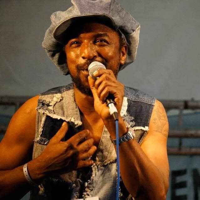 Kareem Shabazz singing with a hand on his chest in a patchwork denim waistcoat on an outdoor festival stage